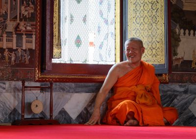 Smiling Buddhist monk in saffron robes seated on a red carpet inside Wat Pho temple in Bangkok, Thailand, bathed in soft daylight from a nearby window, surrounded by traditional murals.
